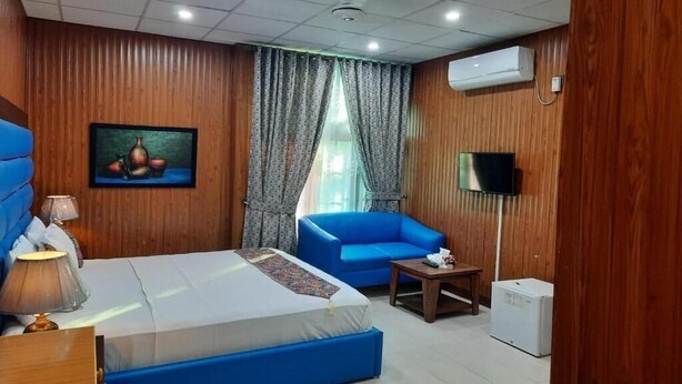 A hotel room with a bed, a blue sofa, a small table, a TV, and an air conditioning unit. - Royal Motel Executive (Islamabad)