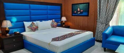 A bedroom with a large bed, wooden panel walls, a painting, and a blue chair.
