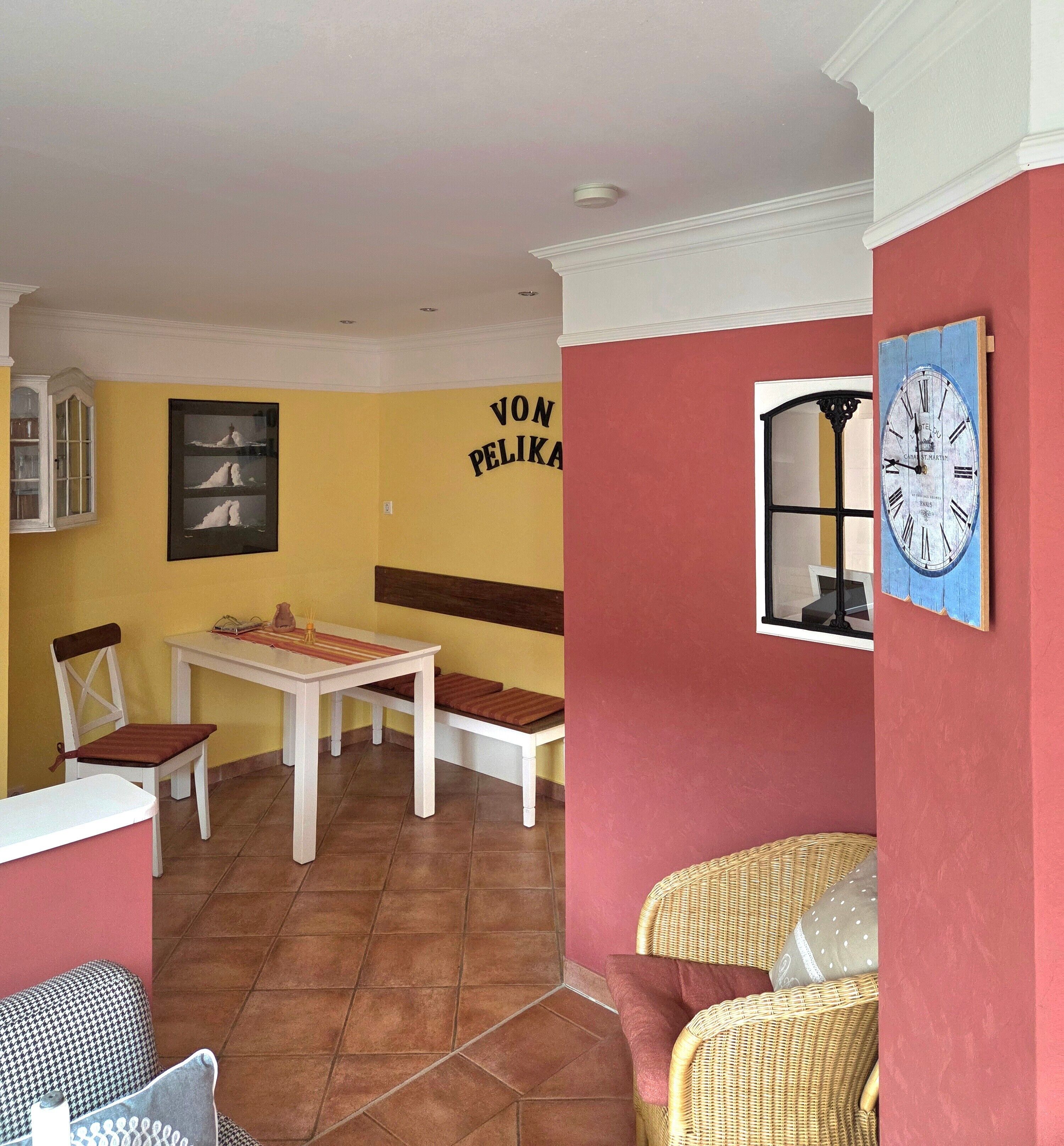 A dining area with a white table and chairs, a wicker chair, a clock on the wall, and a cabinet with glass doors.