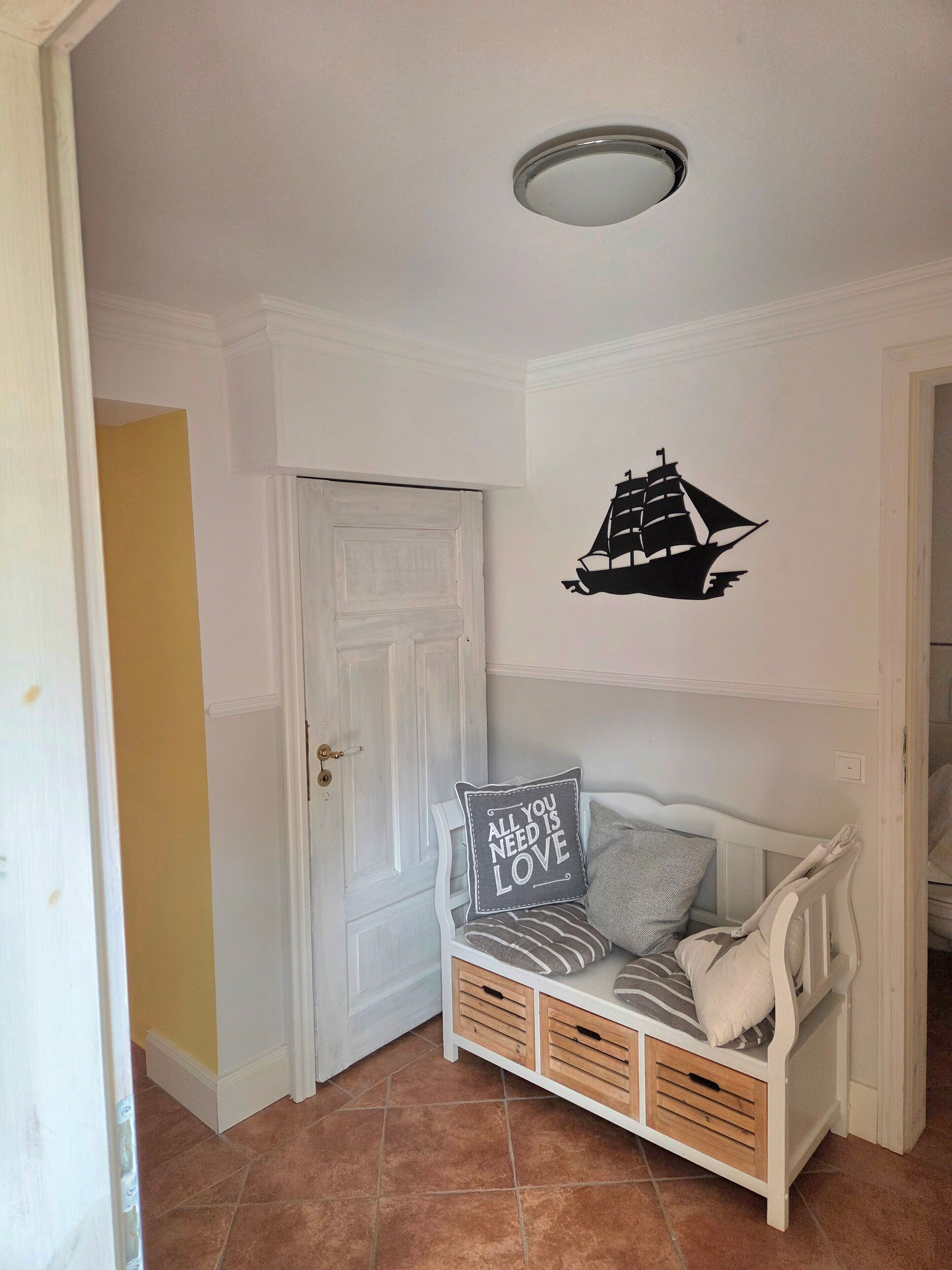 A room with a white door, a wall-mounted ship decal, a bench with pillows, and a ceiling light.