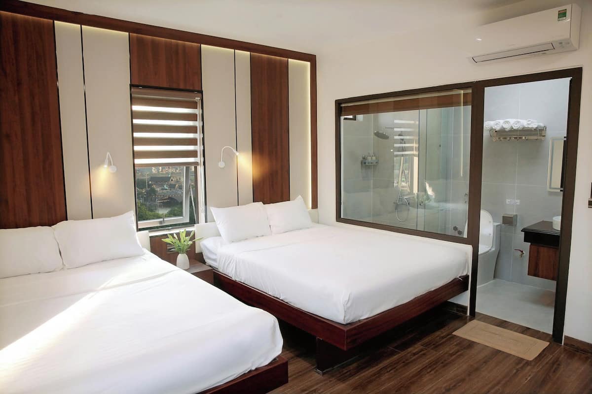 A hotel room with two beds, a large window, a bathroom with a glass shower, and a view of the city.