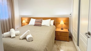 A hotel room with a bed, two bedside tables with lamps, a wooden headboard, a door, and a window with curtains. - Suite Marina Sol (Casares Costa)