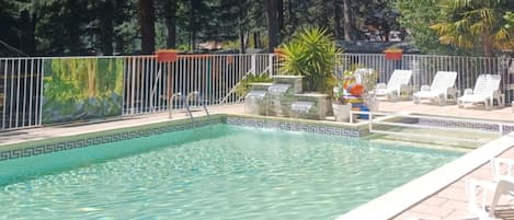 A rectangular pool with a decorative border, surrounded by a white fence. There are lounge chairs and a small garden area with plants and a colorful sculpture.