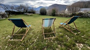 Three wooden deck chairs with blue cushions in a grassy yard with a view of mountains and a lake.