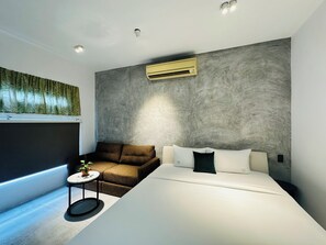 Ein modernes Hotelzimmer mit einem großen Bett, einer Couch, einem kleinen runden Tisch mit Pflanze und einer Klimaanlage an der Wand.