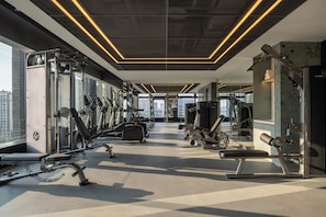 A modern gym with various exercise equipment, large windows, and a high ceiling.