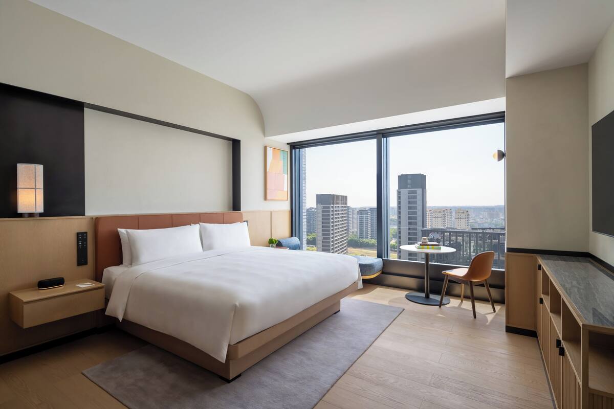 Habitación de hotel moderna con una cama grande, mesita de noche, lámpara y vistas de la ciudad a través de un ventanal amplio.