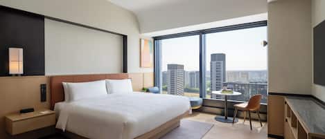 Een moderne hotelkamer met een groot bed, nachtkastje, lamp en uitzicht op de stad door een groot raam.