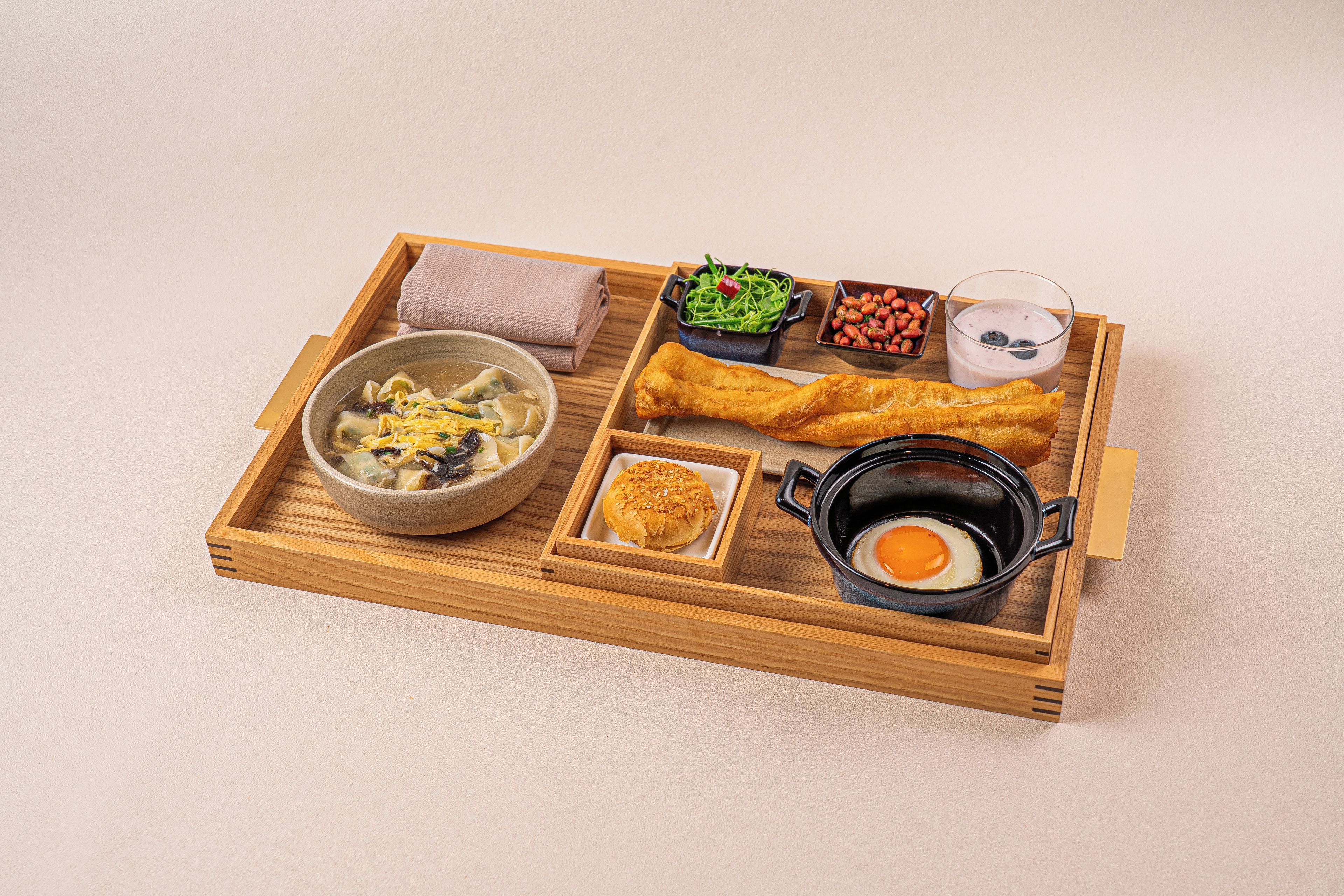 A wooden tray with a bowl of soup, a fried egg, a piece of bread, a small bowl of greens, a bowl of strawberries, and a glass of milk.