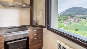 A kitchen with a stove, oven, and wooden cabinets, a window with a view of a village and hills. - Villa Camila 6 - La Panera (Villarraba)