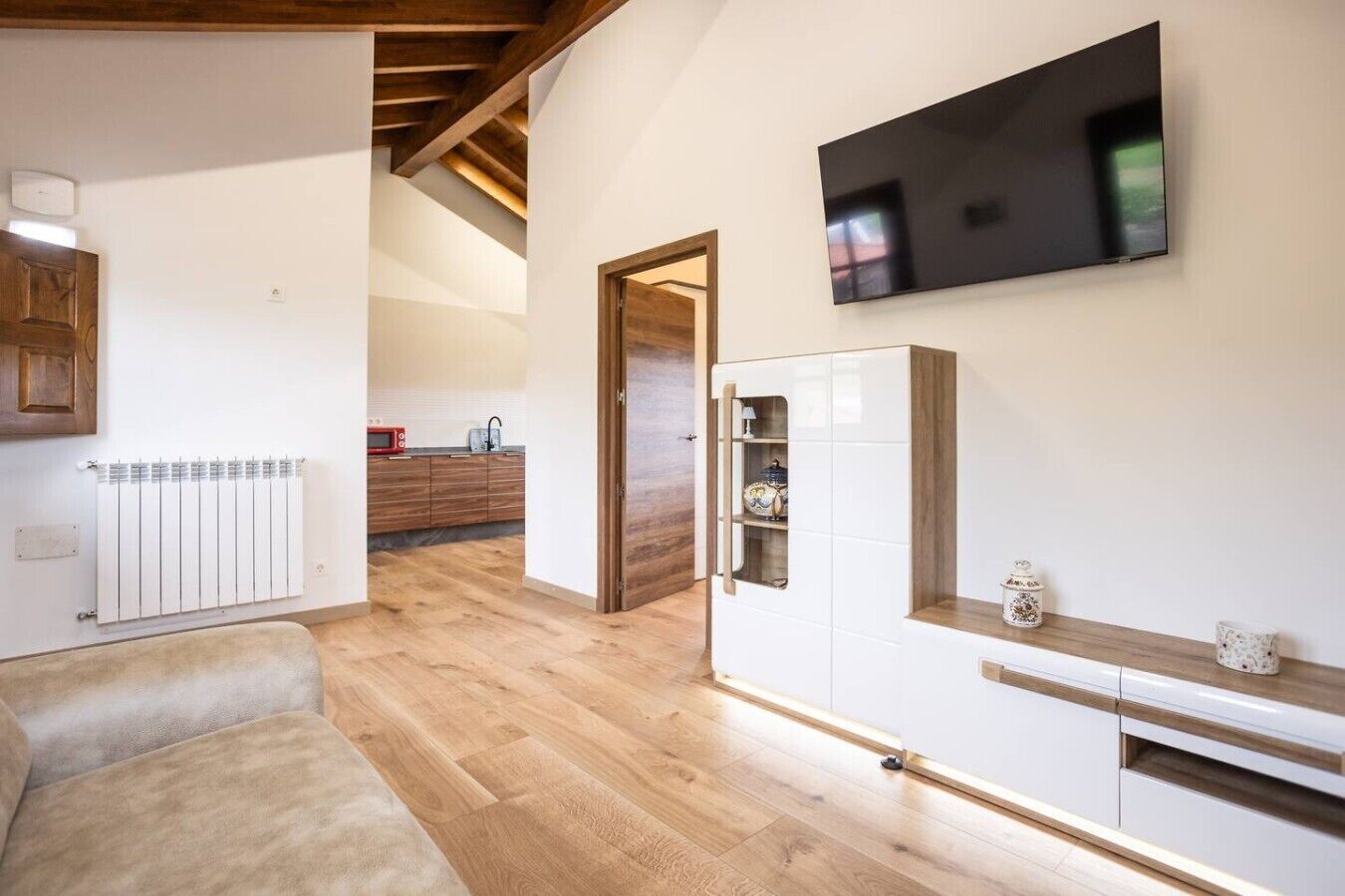 Un salon moderne avec un canapé, un téléviseur fixé au mur, un coin cuisine à l’arrière-plan et un plafond en bois.