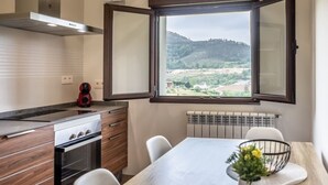 A compact kitchen with a stove, oven, and a coffee maker. A dining table with chairs is positioned under a window with a view of a hilly landscape. - Villa Camila 6 - La Panera (Villarraba)