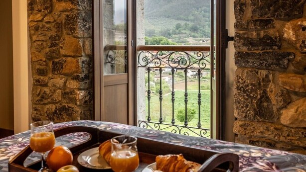 Un mur de pierre, un balcon orné d’une balustrade en fer forgé et une table avec des aliments pour le petit-déjeuner.