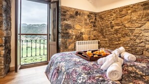 Une chambre avec un mur en pierre, un lit recouvert d’une courtepointe à motifs floraux, un plateau avec des articles pour le petit-déjeuner et une vue sur la campagne, visible à travers une porte vitrée.