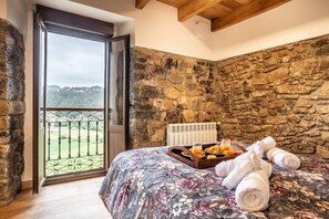 A bedroom with a stone wall, a bed with a floral bedspread, a tray with breakfast items, and a view of the countryside through a glass door.