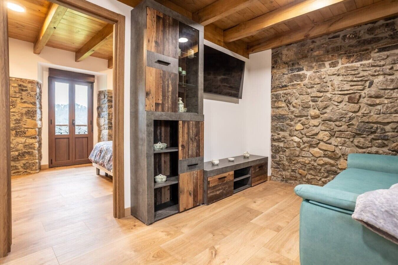Ein rustikaler Raum mit Steinwand, Holzbalken, einem Bett, einem an der Wand montierten Fernseher und einer Couch.