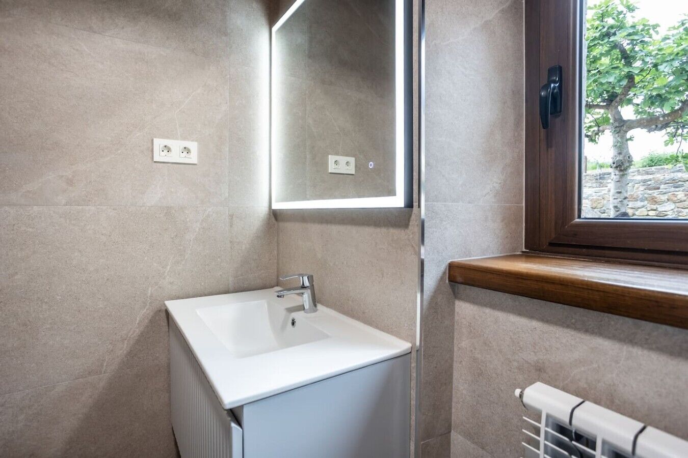Un baño con lavamanos blanco, un espejo, una ventana con vistas a árboles y un radiador.