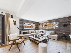 A modern living room with a sofa, a coffee table, and a chair. There are two windows with views of buildings. - Central apartment, spa access - 10 guests (Les Gets)