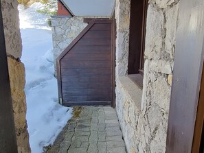 Ein schmaler Steinweg führt zu einer dunklen Holztür, bedeckt mit Schnee.