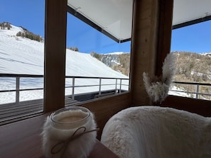 Ein Zimmer mit großem Fenster, das auf eine verschneite Landschaft blickt, einem Holztisch mit einem Glasbehälter und einem weißen, flauschigen Gegenstand.