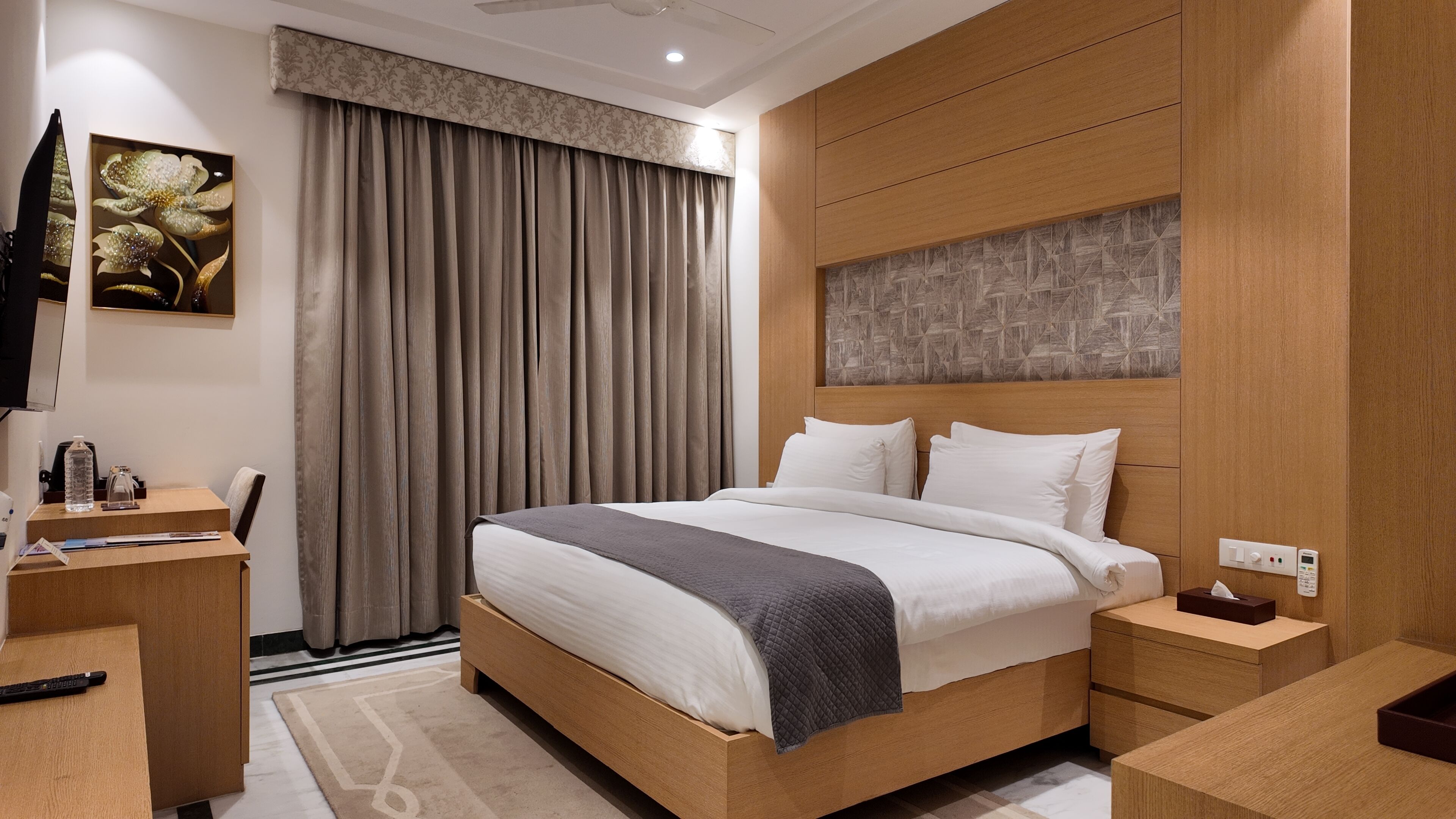 A modern hotel room with a wooden bed, a desk, a chair, a flat-screen TV, and a large window with curtains.