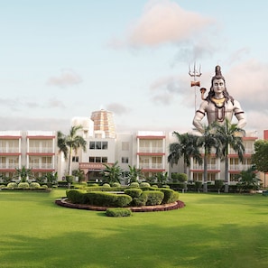 A large statue of a deity in a landscaped area with a hotel building in the background. - Annekaa Heights (Vrindavan)