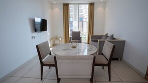 Un salón moderno con una mesa redonda de mármol, dos sofás, un televisor montado en la pared y un ventanal con cortinas.