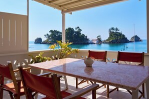 A covered outdoor seating area with a table and chairs, overlooking a coastal landscape with trees and rocks. - 1-bedroom apartment in the heart of Parga (Parga)