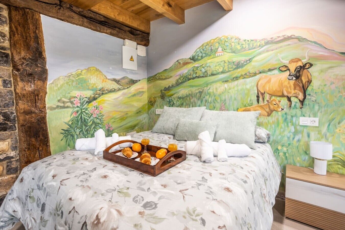 A bedroom with a mural of a pastoral scene, a bed with floral bedding, and a tray of breakfast items.