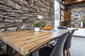 Una mesa de madera para comer con una planta en maceta y un frutero, frente a una pared de piedra y con una cocina al fondo.