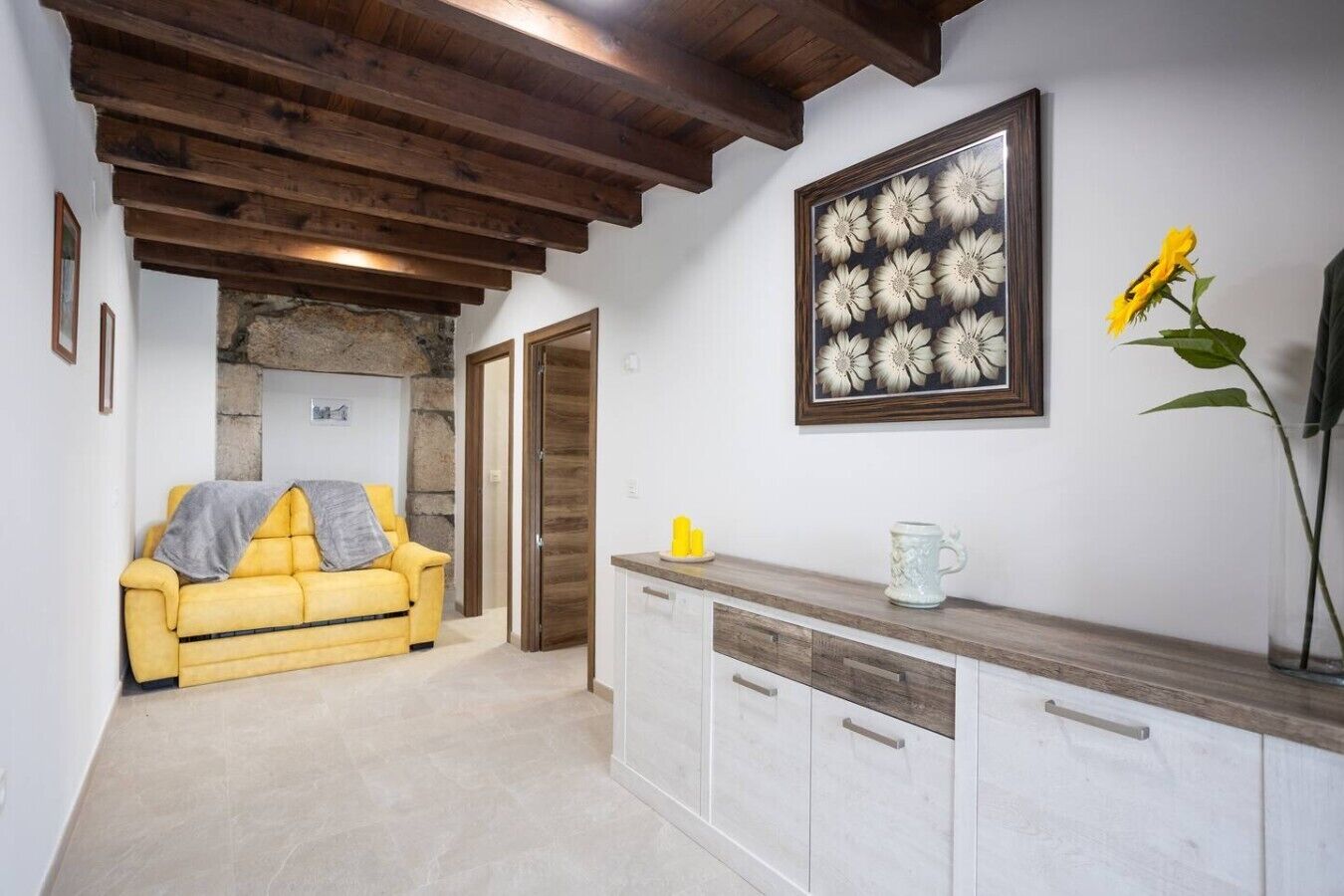 A room with a yellow sofa, a stone wall, wooden beams, a cabinet, and a framed floral artwork.