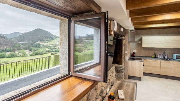 A kitchen with a stone countertop, a window with a view of a green landscape, and wooden cabinets. - Villa Camila - El llagar (Villarraba)