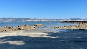 Una playa costera con terreno rocoso, cielo azul despejado y un paisaje urbano lejano.