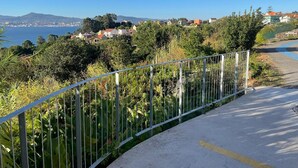 Una carretera costera con una barandilla de metal, un camino pavimentado y vistas a casas y montañas en la distancia.