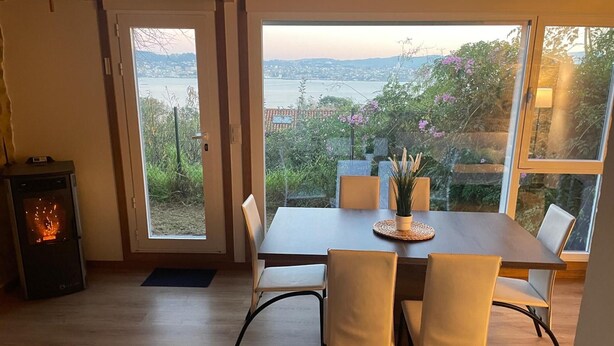 Un área de comedor con una mesa y sillas, chimenea y ventanas amplias con vistas a zonas verdes y una masa de agua.
