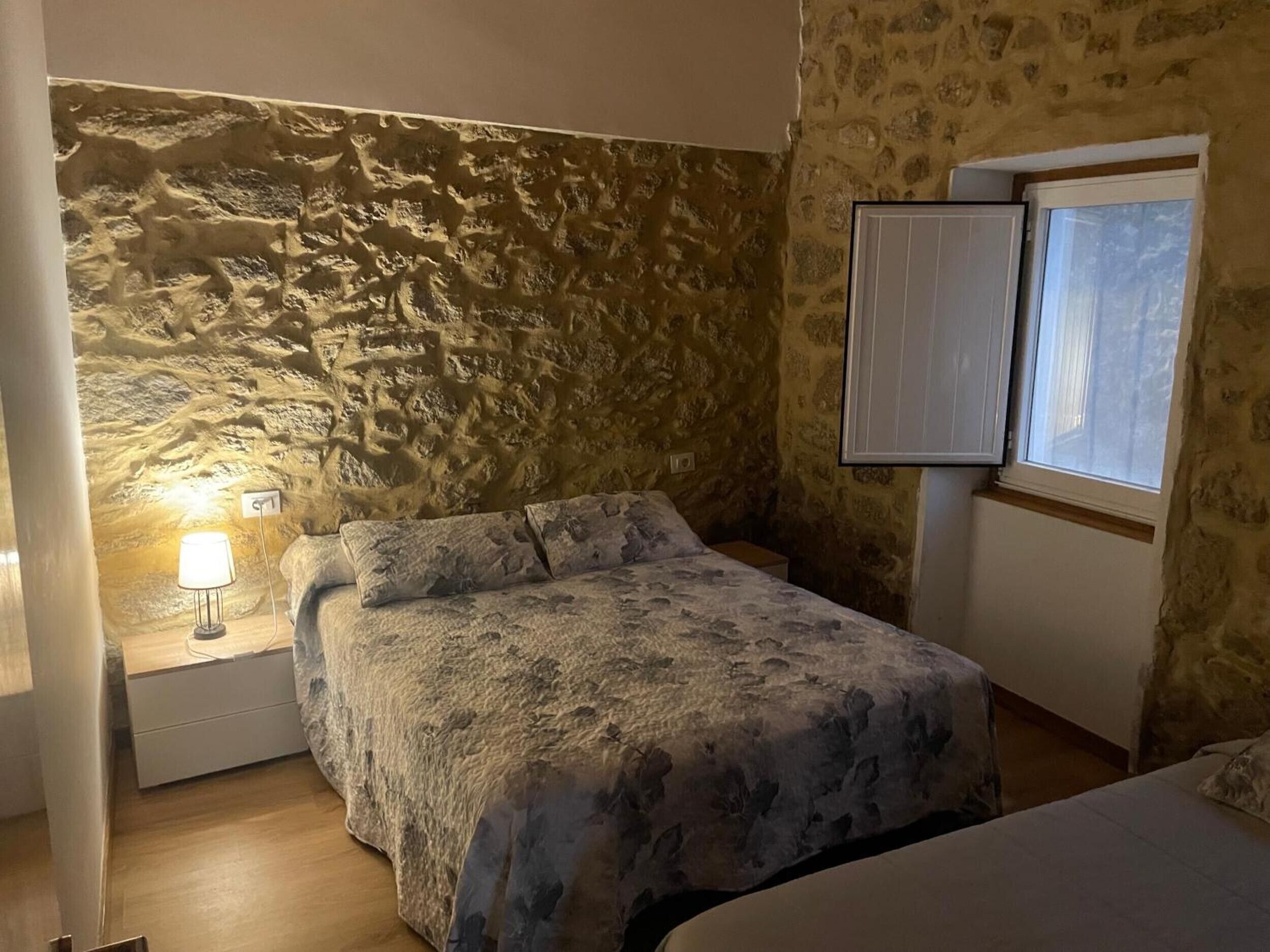 Een slaapkamer met een stenen muur, een bed met een bloemenbedekking, een bedlampje en een raam met jaloezieën.