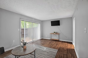 A room with a large sliding glass door, a flat-screen TV mounted on the wall, and a small wooden side table. - Spacious 2BR with 1.5 baths, modern living (Riverdale)
