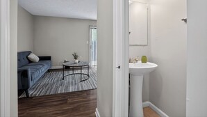 A room with a sofa, a small table with a plant, a white sink, and a door leading to another room. - Spacious 2BR with 1.5 baths, modern living (Riverdale)