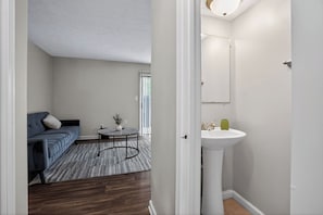 A room with a sofa, a small table with a plant, a white sink, and a door leading to another room. - Spacious 2BR with 1.5 baths, modern living (Riverdale)