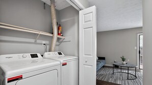 A laundry area with a Whirlpool washer and dryer, a ceiling vent, and a door leading to a bedroom with a bed and a small table. - Spacious 2BR with 1.5 baths, modern living (Riverdale)