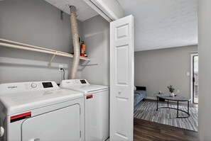 A laundry area with a Whirlpool washer and dryer, a ceiling vent, and a door leading to a bedroom with a bed and a small table. - Spacious 2BR with 1.5 baths, modern living (Riverdale)