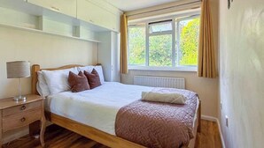 A bedroom with a wooden bed, a nightstand, a lamp, and a window with curtains. - House Sleeps 5 - Wifi & Tv (Luton)