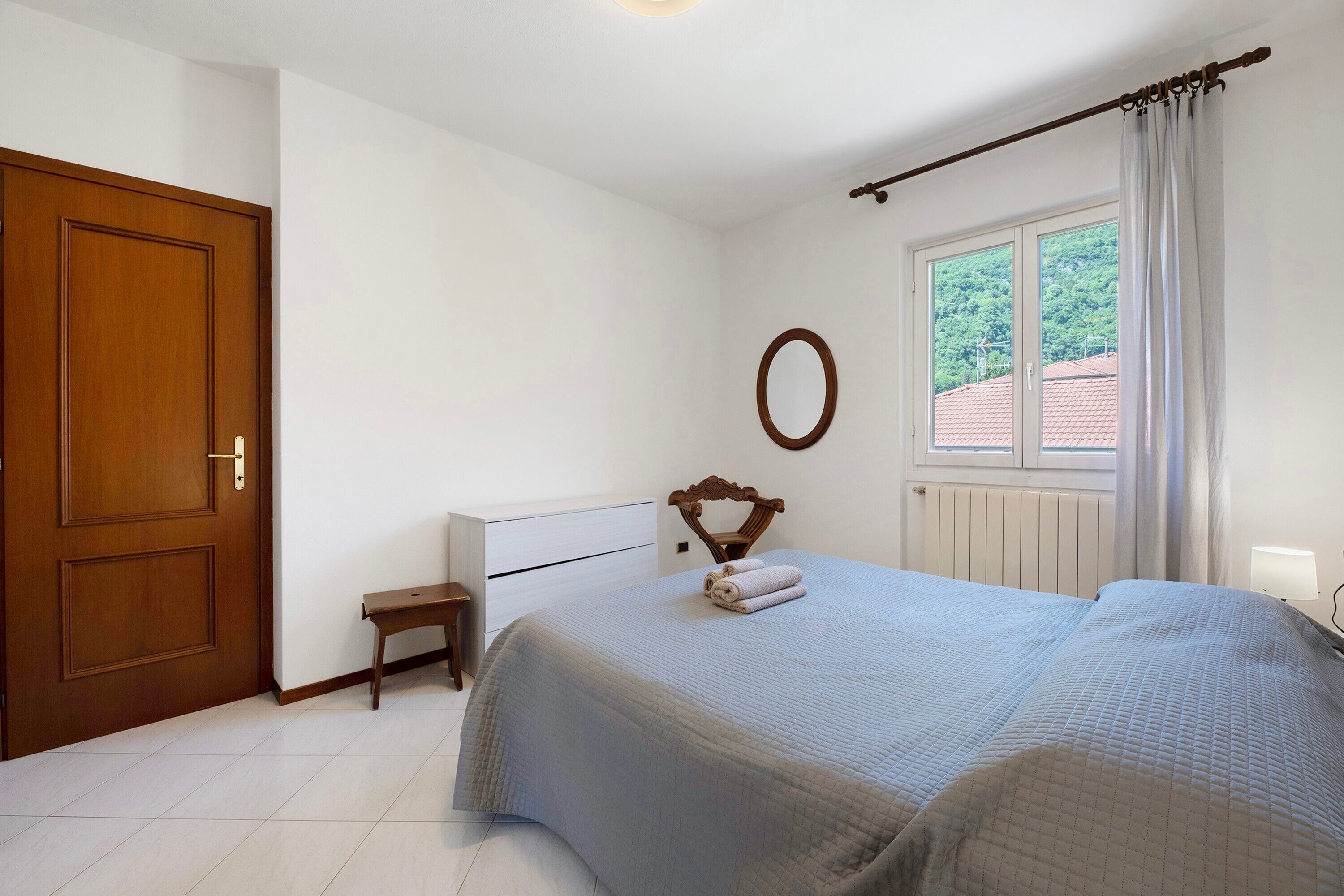 Ein Schlafzimmer mit einem Bett, einer Holztür, einem kleinen Nachttisch, einem Fenster mit Vorhängen und einem an der Wand befestigten Spiegel.