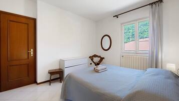 Ein Schlafzimmer mit einem Bett, einer Holztür, einem kleinen Nachttisch, einem Fenster mit Vorhängen und einem an der Wand befestigten Spiegel.