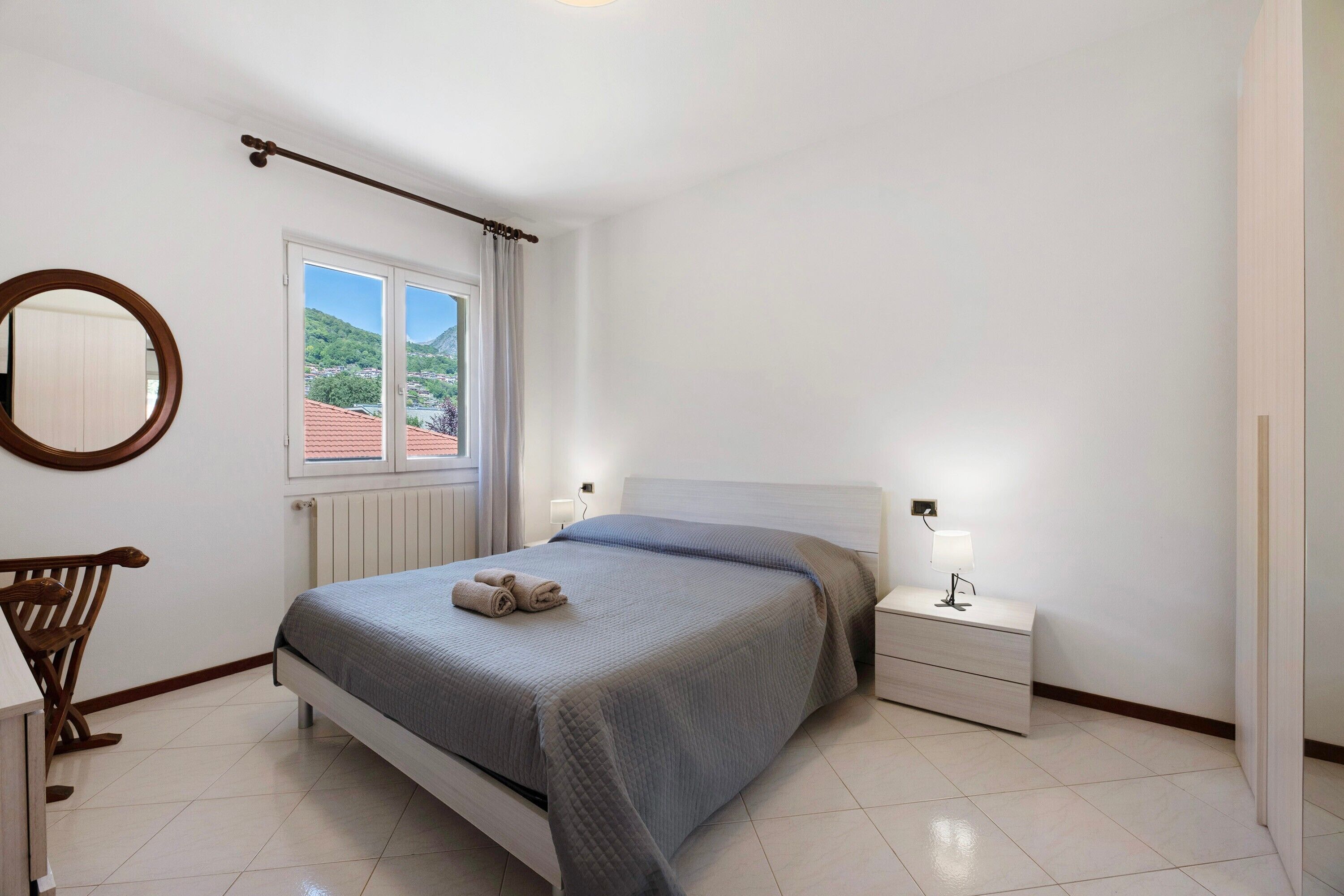 A bedroom with a bed, bedside table, lamp, window, and a view of the outdoors.
