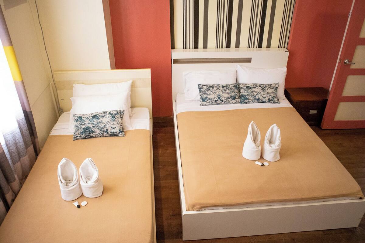 Two single beds with white and patterned pillows, a beige bedspread, and white slippers on each bed.