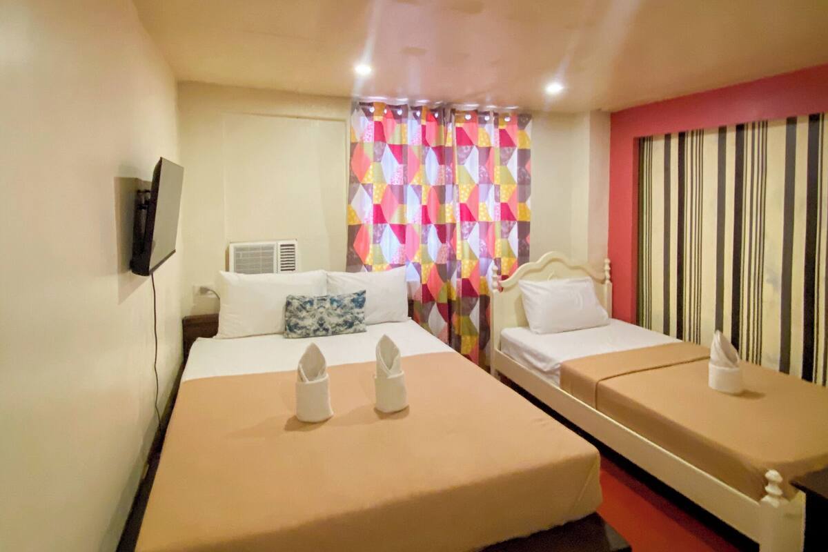 A hotel room with two beds, a TV, an air conditioning unit, and a colorful curtain.