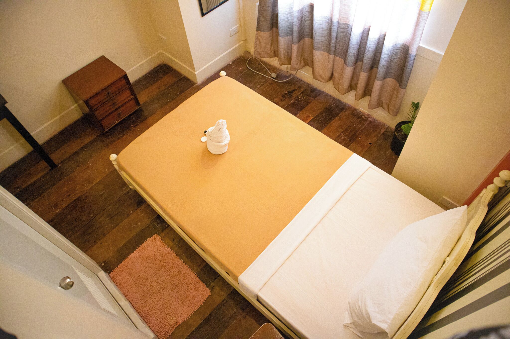 Una habitación pequeña con cama individual, piso de madera, un cofre de madera, un tapete rosa y una ventana con cortinas.