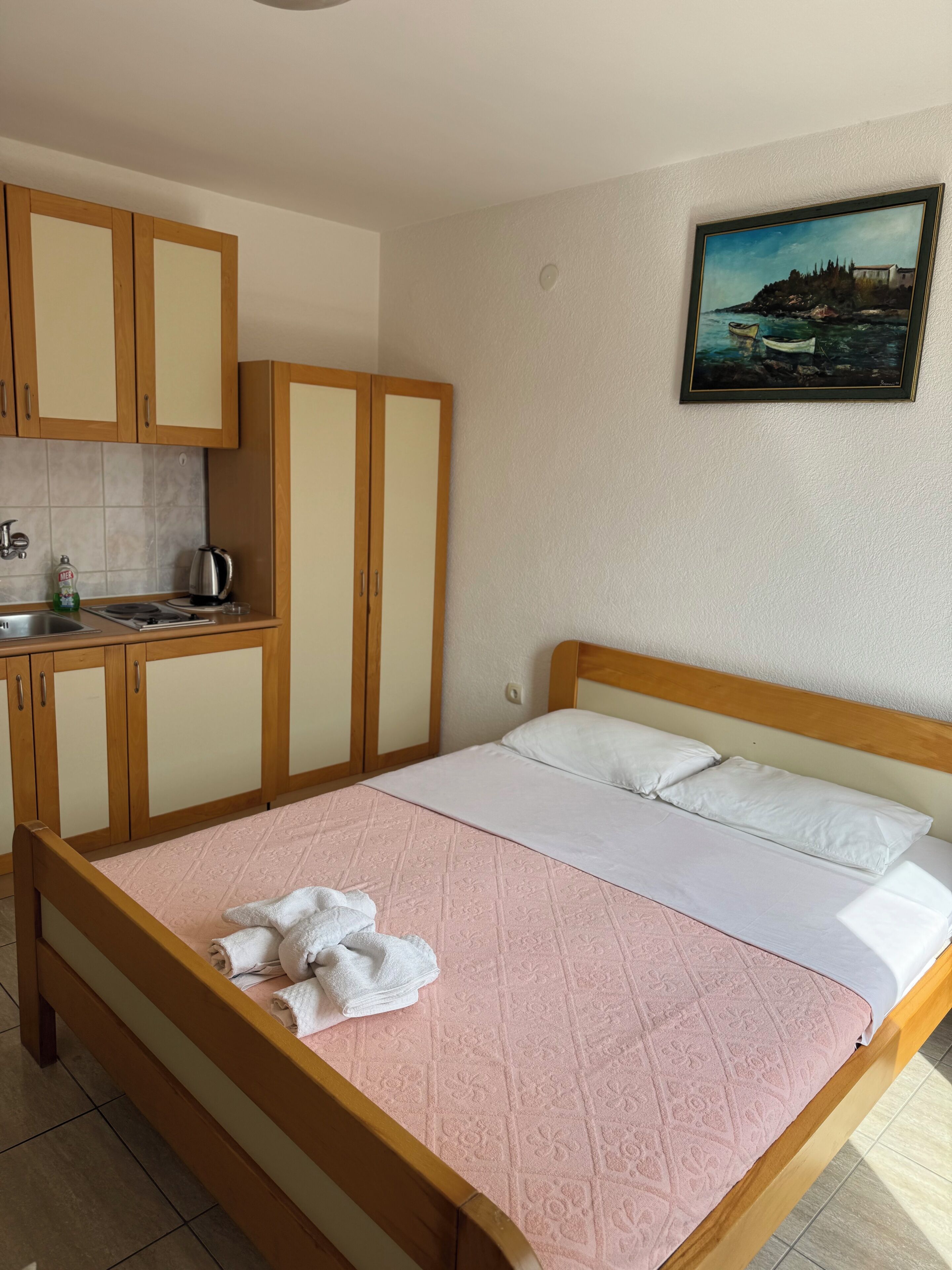 A bedroom with a bed, pillows, a painting on the wall, and a kitchenette with cabinets and a kettle.