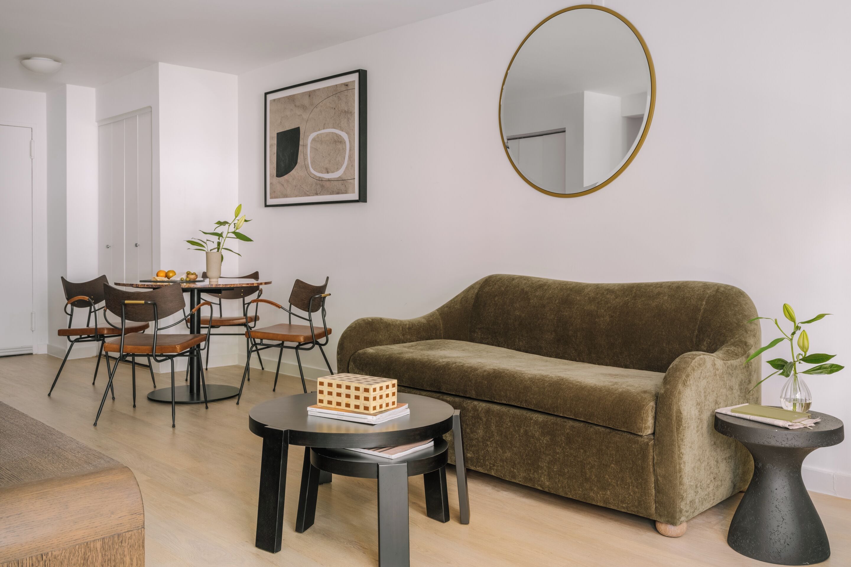 A living room with a brown sofa, a round coffee table, a dining table with chairs, a small side table with a plant, and a framed abstract artwork on the wall.